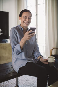 Happy professional with cup using mobile phone while sitting on table in home office