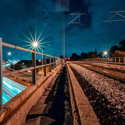 Illuminated railroad tracks by street against sky at night