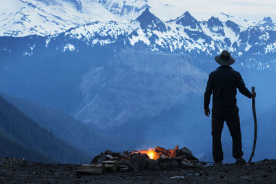 Man holding stick by campfire against snowcapped mountains