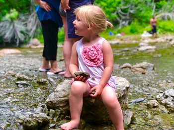 Full length of cute girl looking away while sitting on rock by stream