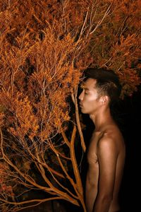 Side view of shirtless young man standing by plant at night