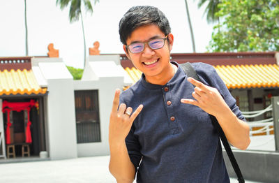 Portrait of happy young man gesturing horn sign while standing against building