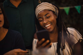 Young woman laughing while looking at mobile phone during dinner party with friends in backyard