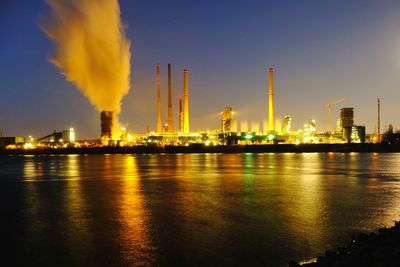 Illuminated factory by river against sky