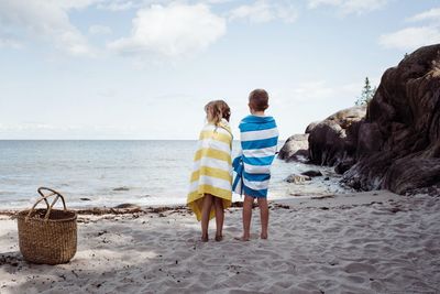 Boy and girl stood on the beach wrapped in towels looking at the ocean