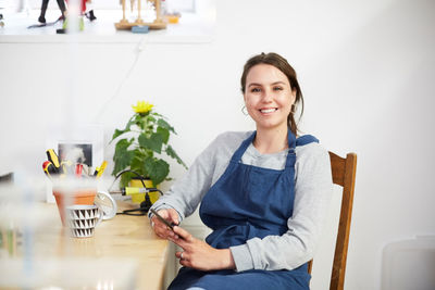 Portrait of smiling young female engineer with smart phone sitting at table in workshop