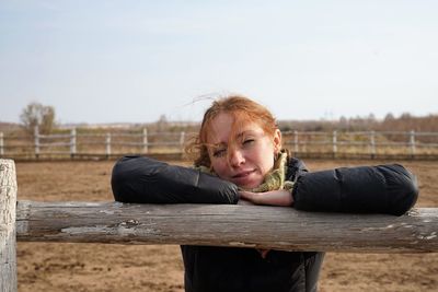 Portrait of woman leaning on wooden fence against sky