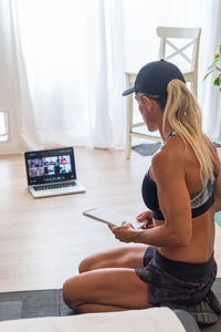 From above of sportswoman in activewear sitting on floor with laptop and reading list of exercises for workout