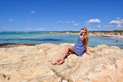 Woman sitting on rock at beach against blue sky