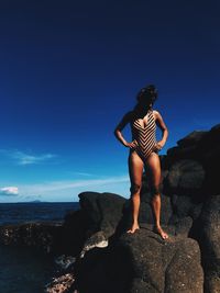 Woman wearing swimsuit while standing on rock against blue sky