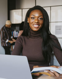 Portrait of smiling young female student sitting with book studying at community college