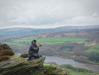 Man holding camera while kneeling on cliff against cloudy sky during foggy weather