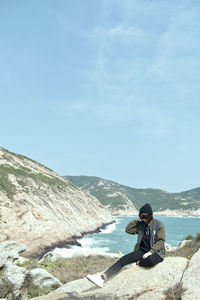 Man sitting on mountain by sea against sky