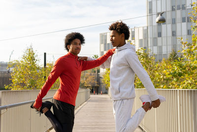 Side view of african american males doing stretching legs while leaning on each other on street near fence