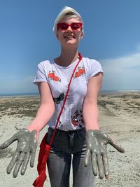 Portrait of young woman wearing sunglasses showing messy hands at beach against sky