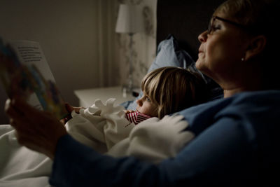 Senior woman reading story book for grandson on bed at home