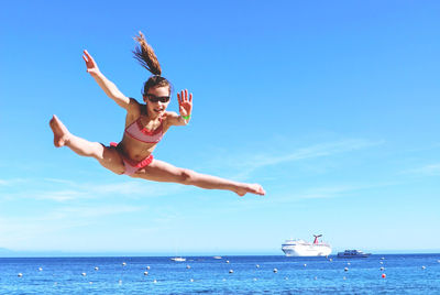Portrait of girl doing straddle jump on beach against blue sky
