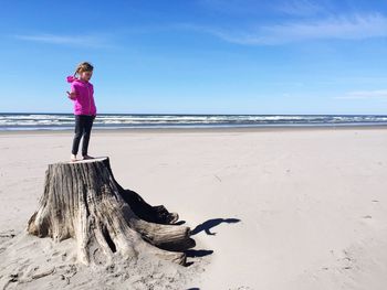Full length of girl standing on tree stump at beach against sky