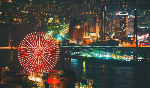 Illuminated ferris wheel by buildings against sky at night