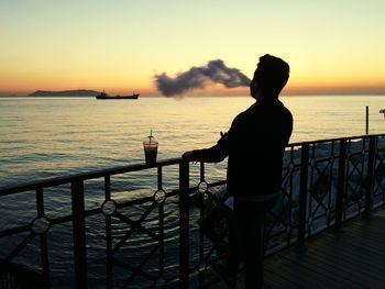 Silhouette young man looking at sea while standing by railing against sky during sunset