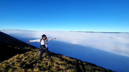 Front view of a man, with arms wide open, on top a mountain and a sea of clouds in the background. 