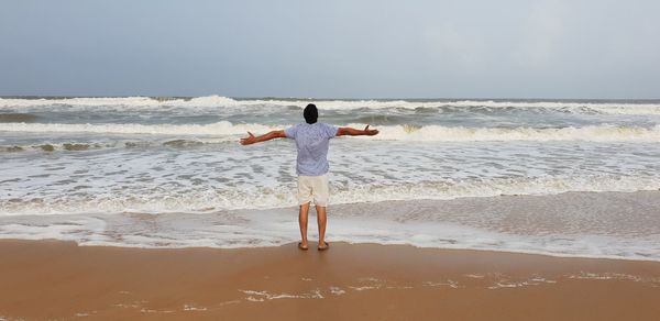 Rear view of man with arms outstretched standing on shore at beach against sky
