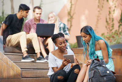 Young friends using laptop and tablet sitting on steps