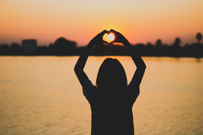 Silhouette woman making heart shape with hands by lake against sky during sunset