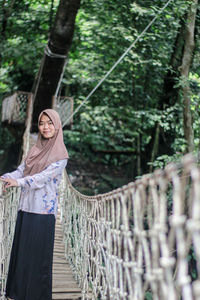 Portrait of woman wearing hijab standing on rope bridge against trees in forest