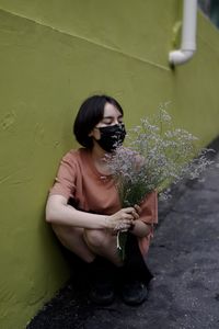 Young woman holding flower while standing against wall