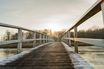 Footbridge over frozen river against sky during winter