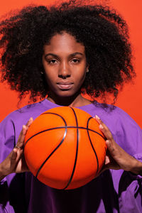 Emotionless ethnic woman with afro hair wearing purple sport shirt and holding bright orange basketball in studio