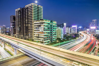 Light trails on road by buildings against sky at night