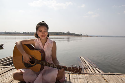 Portrait of smiling young woman with guitar sitting on pier over lake against sky