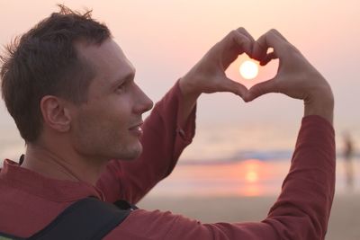 Side view of mid adult man making heart shape against sky during sunset