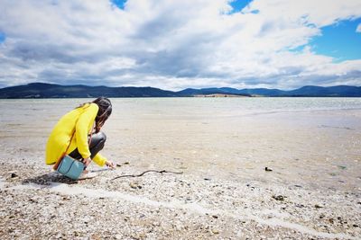 Side view full length of woman crouching at riverbank against sky