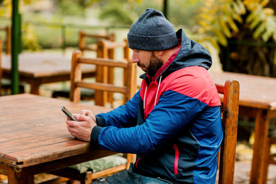 A young man using a mobile phone is sitting outdoors in a cafe or restaurant on an autumn day