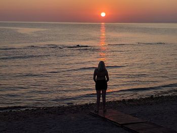 Full length rear view of woman standing at beach against sky during sunset