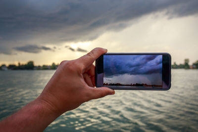 Cropped hand of man photographing cloudy sky from smart phone during sunset