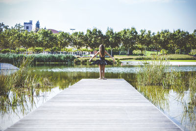 Rear view of woman in ballerina costume standing on pier over lake against sky