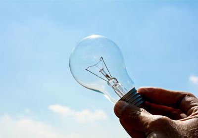 Low angle view of hand holding light bulb against sky