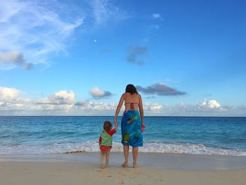 Full length rear view of mother with daughter standing at beach against sky