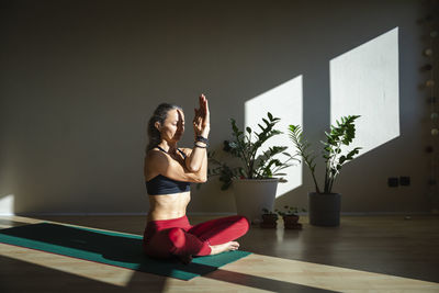 Mature woman with cross legged practicing yoga on mat at home during sunny day