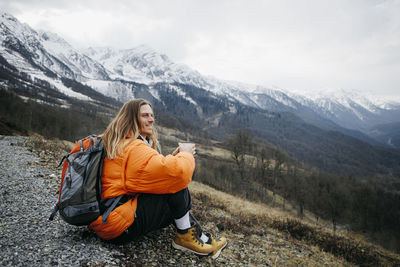 Smiling man with coffee mug and backpack sitting on mountain