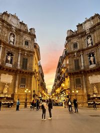 People walking on street amidst buildings in city at sunset