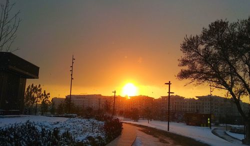 Snow covered houses by trees against sky during sunset