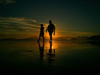 Silhouette man and woman walking on shore against sky during sunset