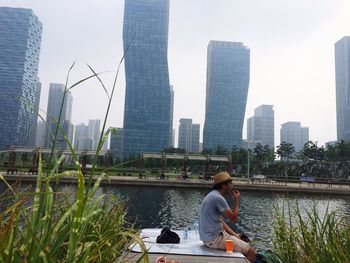 Side view of man sitting on jetty in river against skyscrapers