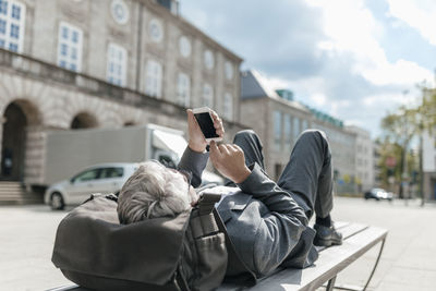 Senior businessman using smartphone, lying on bench in the city