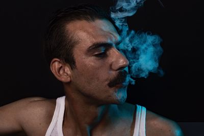 Close-up of young man smoking while standing against black background